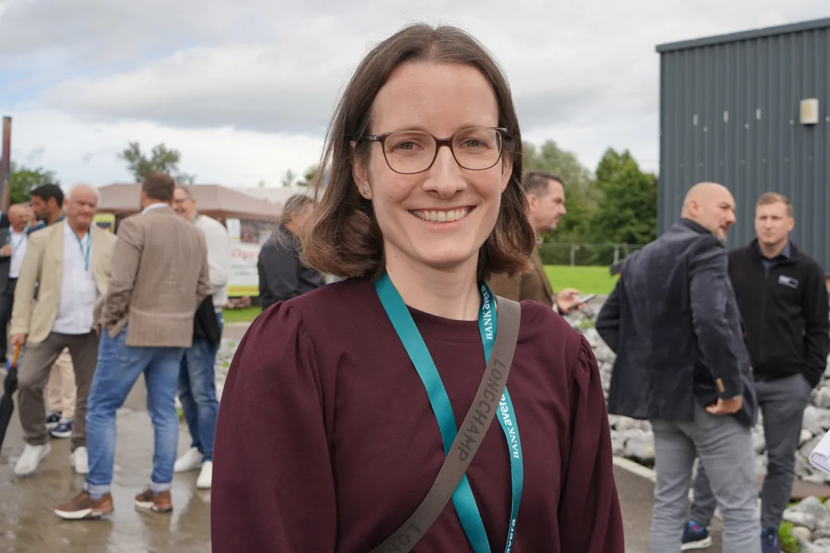 Eine dunkelhaarige Frau mit weinroten Pullover. Man sieht den Gurt einer Umhängetasche über ihrer Schulter.